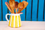 A white ceramic pitcher with bold yellow vertical stripes, filled with several wooden kitchen utensils including spoons and spatulas, set on a light wood surface against a bright blue wooden slat background.