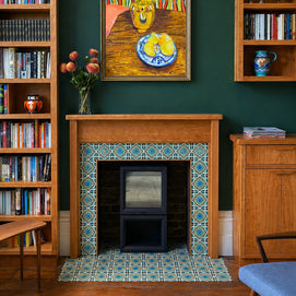 A styled interior scene showing a wood-burning fireplace with a blue and white geometric tile surround. The room has dark green walls, wooden bookshelves filled with books, a framed still life painting above the fireplace, and a small cabinet to the right.