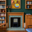 A styled interior scene showing a wood-burning fireplace with a blue and white geometric tile surround. The room has dark green walls, wooden bookshelves filled with books, a framed still life painting above the fireplace, and a small cabinet to the right.
