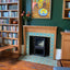 A styled interior scene showing a wooden fireplace with turquoise and cream patterned tiles, a black wood-burning stove insert, a bookshelf, and wall art in a real room environment.