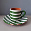 A handmade ceramic coffee cup and saucer set with green and white stripes and a terracotta orange interior, displayed on a light gray wooden surface against a dark gray background.
