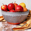 A ceramic bowl filled with red and yellow apples, placed on a rustic wooden surface with a folded tan cloth underneath. The bowl has a speckled gray and orange glaze.