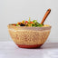 A handmade ceramic bowl with a speckled beige and terracotta pattern, filled with a colorful salad and topped with a wooden spoon, set against a neutral light gray background.