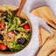 A bowl of mixed salad with greens, cherry tomatoes, shredded carrots, and dark beets, served with a wooden spoon on a light gray surface. A folded tan cloth napkin with black lace trim sits to the right.