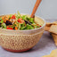 A ceramic salad bowl with a speckled beige pattern and a wooden spoon, filled with a fresh green salad containing tomatoes, carrots, and beets, set on a gray surface with a yellow napkin.