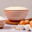 A styled kitchen scene with a large terracotta-and-white striped bowl filled with flour, a smaller matching bowl of flour, brown eggs in a white carton, and a wooden spoon on a light surface against a soft neutral background.