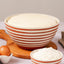 A terracotta-colored ceramic bowl with horizontal white stripes sits on a wooden cutting board, filled with risen dough. A smaller matching bowl in front holds flour, with eggs, a wooden spoon, and a beige cloth arranged around it on a light stone surface against a soft neutral background.