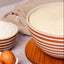 A large ceramic bowl with terracotta and cream horizontal stripes, filled with risen dough, sits on a wooden board. A smaller matching striped bowl contains white flour, next to a white egg carton with three brown eggs and a small wooden spoon on a light purple surface.