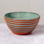 A handmade ceramic bowl with horizontal orange and sage green striped patterns, set against a neutral light gray background.