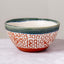 A handmade ceramic bowl with a white base decorated with a red speckled pattern, a dark green glazed rim, and a solid red foot ring, set against a neutral gray background.
