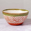 A handmade ceramic bowl with a white base decorated with a red leaf pattern, a green glazed rim, and a terracotta-colored bottom, set against a neutral gray background.