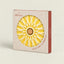C. Atlántica hot pot stand in its original packaging, featuring a yellow and white starburst design with a cork center, set against a light beige background.