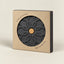 A dark gray ceramic pot base with a raised floral pattern and a small cork center, shown inside a kraft paper box with a circular cutout. Text on the box reads 'FERAMIC POT BASE' and 'MADE IN PORTUGAL'.