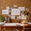 A styled interior workspace with a corkboard wall covered in architectural drawings, notes, and small photos, above a wooden desk with blueprints, books, a lamp, and decor.
