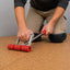 A person kneels on a tan cork floor while using a red and silver floor roller tool to smooth or level the cork surface.