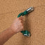A close-up of a person's hand holding a green and silver wall-mounted grab bar against a cork tile wall.