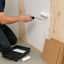 A person kneels on a concrete floor while painting a wall with a white roller. A black paint tray with a paint sponge, a small spare roller, and stacked cork boards are visible on the floor.
