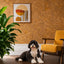 A black and white dog lies on a dark wood floor in a cozy room with a textured cork wall, a yellow armchair, a green houseplant, and a framed abstract art print.
