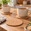 Two beige speckled ceramic mugs filled with tea sit on round cork coasters on a wooden table, with a third coaster in the foreground and a folded striped cloth beside them.