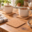 Two beige speckled ceramic mugs sit on cork coasters on a wooden table, with a folded striped cloth and a bowl of cookies nearby. The background shows a softly blurred kitchen scene with plants and teapots.