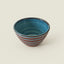 A small ceramic bowl with a glossy blue interior and horizontal reddish-brown and blue stripes on the exterior, set against a plain light beige background.
