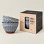 Two small ceramic bowls with a speckled blue and brown glaze, shown next to their black and kraft paper packaging box on a neutral beige background.