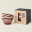 Two small striped ceramic bowls in a stacked arrangement next to their kraft paper and black cardboard packaging box, set against a solid light beige background.