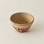 A small handmade ceramic bowl with a speckled brown pattern on a beige exterior and a smooth tan interior, set against a plain light beige background.