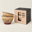 Three small speckled ceramic bowls with a brown base are shown next to their black and kraft paper packaging box on a light beige background.