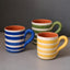 Three ceramic mugs with horizontal stripes in blue, green, and yellow, featuring terracotta orange interiors, arranged on a gray surface against a dark gray background.