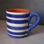 A ceramic mug with blue and white horizontal stripes, featuring a textured blue handle and a contrasting orange interior, set against a neutral gray background.