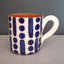 A ceramic mug with a white exterior decorated with blue painted vertical stripes and dots, featuring a terracotta orange interior and a white handle, set against a dark gray background.