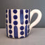 A white ceramic mug with a blue hand-painted abstract pattern of vertical stripes and dots, set against a neutral gray background.