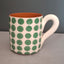 A ceramic mug with a white exterior decorated with green polka dots, featuring a terracotta orange interior and a white handle, set against a dark gray background.