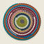 A round hand-crocheted rug with concentric rings of multicolor yarn, including teal, orange, burgundy, pink, olive green, navy, white, and gray, on a light beige background.