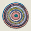 A round braided rope mat with concentric rings of blue, pink, green, gray, and white yarn, photographed flat against a light beige background.