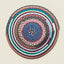 A round braided rug with concentric rings of pink, blue, teal, gray, brown, and white yarn, photographed flat against a light beige background.