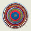 A round hand-crocheted rug with concentric rings of blue, red, green, brown, orange, and white yarn, featuring a small woven symbol in the center, set against a solid light beige background.