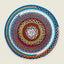 A round braided placemat with concentric rings of multicolor yarn, photographed flat against a light beige background.