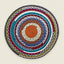 A round, handwoven braided placemat with concentric circular patterns in multiple colors including orange, teal, blue, burgundy, olive green, and white, set against a solid light beige background.