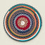 A round, hand-crocheted braided placemat with concentric rings of red, navy, teal, orange, olive green, and white yarn, centered on a light beige background.