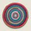 A round, hand-crocheted woven placemat with concentric rings of multiple colors including pink, teal, blue, red, brown, and gray, centered on a light beige background.