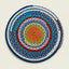 A round braided rug with concentric rings of blue, teal, red, olive green, yellow, and orange yarn, photographed flat against a light beige background.