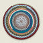 A round braided rope placemat with concentric circular patterns in multiple colors, including red, teal, blue, pink, gray, white, and yellow, photographed flat against a light beige background.