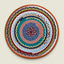 A round braided woven rug or mat with concentric rings of orange, brown, red, blue, teal, pink, white, and olive green, photographed flat against a light beige background.