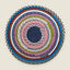 A round braided rug with concentric rings of multiple colors, including blue, pink, green, white, and brown, on a light beige background.