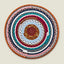 A round hand-crocheted rug with concentric rings of orange, pink, white, navy, teal, burgundy, light blue, and brown yarn, featuring a small woven floral motif in the center, set against a solid light beige background.